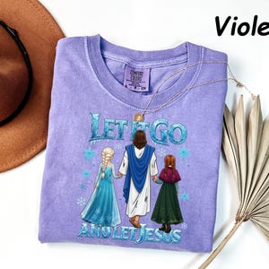 May include: Violet-colored t-shirt with the text "LET IT GO AND LET JESUS" and a graphic of Jesus holding hands with two girls. The shirt has a crew neck and short sleeves. A gold necklace is draped across the shirt.