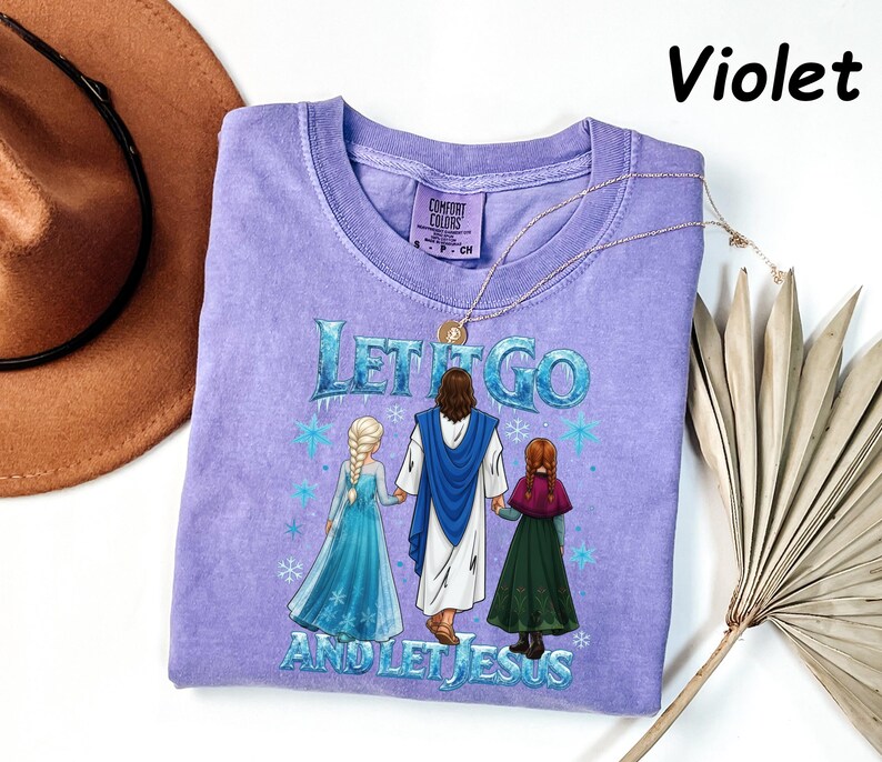 May include: Violet-colored t-shirt with the text "LET IT GO AND LET JESUS" and a graphic of Jesus holding hands with two girls. The shirt has a crew neck and short sleeves. A gold necklace is draped across the shirt.