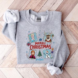 Puede incluir: Una sudadera gris con personajes de la pel&iacute;cula Frozen, incluyendo a Elsa, Anna, Olaf y Sven, con las palabras "Merry Christmas" y "Frozen" impresas en la parte delantera. La sudadera es de un material suave.