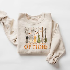 May include: Cream-coloured sweatshirt with a graphic of several witch brooms in different colours and styles. The text "A GIRL'S GOTTA HAVE OPTIONS" is printed in orange below the brooms. The sweatshirt has a crew neck.