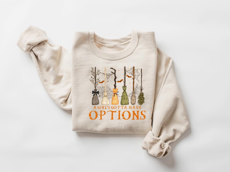 May include: Cream-coloured sweatshirt with a graphic of several witch brooms in different colours and styles. The text "A GIRL'S GOTTA HAVE OPTIONS" is printed in orange below the brooms. The sweatshirt has a crew neck.