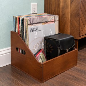 May include: A dark brown wooden storage crate with side handles, filled with a collection of vinyl records and a black leather-like case. The front-most record sleeve features the text "BLA CE" and a handwritten signature in black ink.