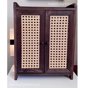 May include: A dark brown wooden cabinet with two doors featuring woven cane panels. The cabinet has a rectangular shape with a small shelf and two round knobs. The text "handybykamran" is visible in the image.