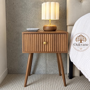 May include: A wooden nightstand with a single drawer featuring a vertical fluted texture and a small round brass knob. The nightstand stands on four tapered wooden legs and is topped with a small lamp with a white pleated shade and a wooden base.