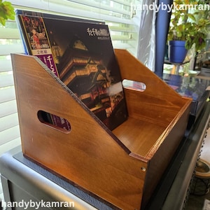 May include: A wooden record holder with a warm brown finish. The holder has a rectangular shape with angled sides and cut-out handles. Two album covers are visible inside the holder, featuring Japanese text and colorful illustrations.