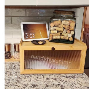 May include: A bamboo bread box with a glass door and a small wooden knob. The bread box is sitting on a granite countertop. A glass jar filled with wine corks is sitting on the counter behind the bread box. A tablet displaying the time 2:32 is sitting on the counter in front of the bread box.