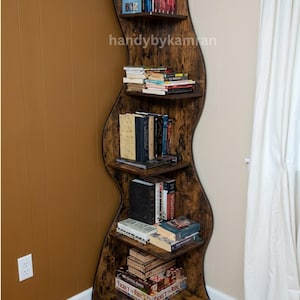 May include: A tall, S-shaped wooden bookshelf with a dark, distressed brown finish. The unit features five horizontal shelves filled with various books and a Ticket to Ride board game box at the base. The text handybykamran is visible.