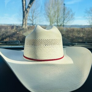 May include: A white cowboy hat with a wide brim and a perforated crown. A thin red band encircles the hat just above the brim. The hat is made of woven material.