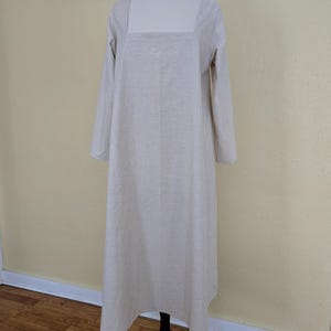 May include: A light beige, long-sleeved dress with a square neckline. The dress is displayed on a white mannequin and has a loose, flowing fit. The dress is floor-length and made of a soft, textured fabric.