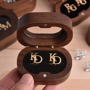 Könnte beinhalten: Eine Holzschmuckschatulle mit offenem Deckel, die goldfarbene Manschettenknöpfe mit den Initialen "KD" auf einem schwarzen Samtinnenraum zeigt. Im Hintergrund sind weitere Schatullen mit verschiedenen Initialen zu sehen. Die Schatulle wird von einer Person gehalten.