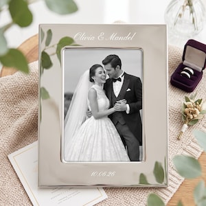 May include: A polished silver-toned rectangular picture frame featuring the engraved text Olivia & Mandel at the top and 10.06.26 at the bottom. The frame holds a black and white photograph of a couple in wedding attire, showing a person in a gown and a person in a suit.