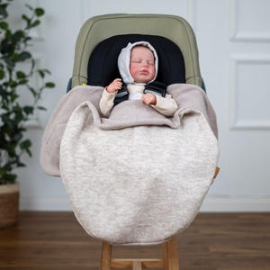 Könnte beinhalten: Ein beiger Fleece-Autositzbezug mit einem schlafenden Baby darin. Das Baby trägt eine weiße Kapuze und ist im Autositz angeschnallt.