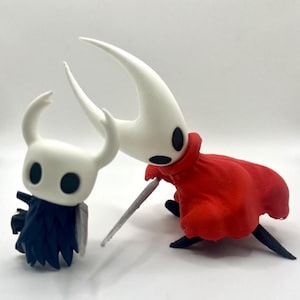 May include: Two figurines of characters from the video game Hollow Knight. One figurine is white with a dark blue cape, and the other is white with a red cape and a sword. The figurines are on a white surface.