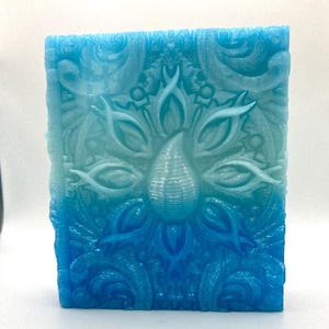 May include: A rectangular bar of soap with a gradient of blue hues. The soap features a detailed, embossed design of a central teardrop shape surrounded by floral and abstract patterns. The soap is approximately 3 inches tall.
