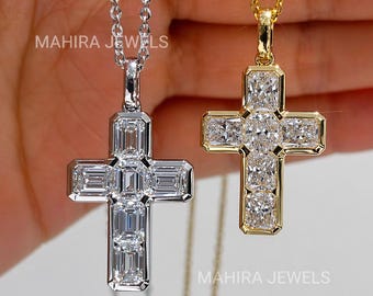 Puede incluir: Dos colgantes de cruz en cadenas, uno en metal plateado y otro en metal dorado. Cada cruz presenta múltiples piedras transparentes rectangulares de corte esmeralda dispuestas en forma de cruz. El texto MAHIRA JEWELS es visible en la imagen.