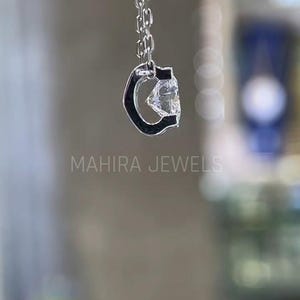 Puede incluir: Un collar de cadena de plata con un colgante de piedra preciosa transparente en forma de letra C. El colgante est&aacute; engastado en un marco de plata.