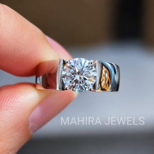 Op de afbeelding: Een zilverkleurige ring met een enkele rondgeslepen heldere edelsteen in een spanningzetting. De ringband is breed en gepolijst met een glad, reflecterend oppervlak. De tekst MAHIRA JEWELS staat in het wit onderaan de afbeelding.