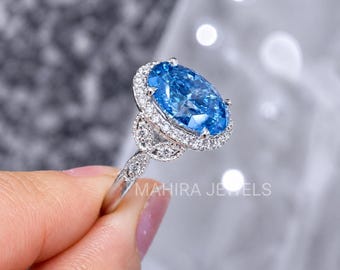 May include: A silver-toned ring featuring a large, oval-cut blue centre stone surrounded by a halo of small, clear, round-cut stones. The band is decorated with additional clear stones in a leaf-like pattern. The text MAHIRA JEWELS is visible.