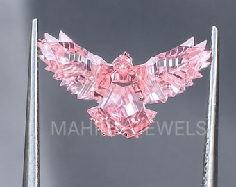 May include: A light pink gemstone cut into the shape of a bird with outstretched wings. The faceted stone displays geometric patterns and sharp edges. The text MAH JEWELS is visible across the center of the gemstone in a light gray font.