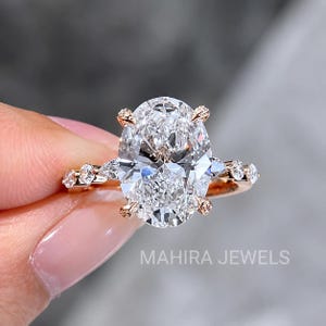Puede incluir: Un anillo de oro rosa con una gran piedra central de corte ovalado sujeta por cuatro garras. La banda está adornada con piedras más pequeñas redondas y en forma de pera a cada lado. El texto MAHIRA JEWELS es visible en la parte inferior.