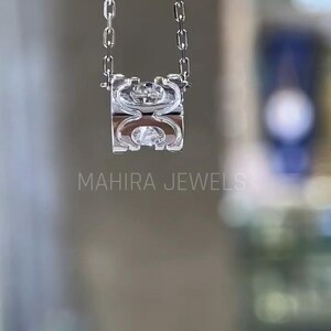 Puede incluir: Un collar de cadena de plata con un colgante de plata que presenta un dise&ntilde;o geom&eacute;trico con un cristal transparente en el centro. El colgante tiene forma de anillo con una abertura cuadrada. El collar est&aacute; colgado sobre un fondo blanco.