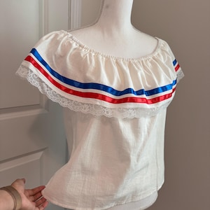 May include: Off-the-shoulder white blouse with red and blue ribbon trim. The blouse features a ruffled neckline with lace edging. The garment is displayed on a mannequin.