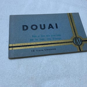 May include: A rectangular, gray booklet with the word "DOUAI" in large, black letters. Gold and black stripes and a circular emblem adorn the top right corner. The text "Paix et bien être pour tous par les Etats-Unis d'Europe" and "12 Vues Choisies" are also visible.