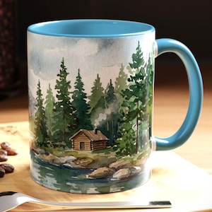May include: A ceramic mug with a light blue interior and handle, featuring a watercolor illustration of a log cabin nestled in a dense pine forest beside a calm lake. The scene includes green trees, rocky shores, and a small chimney with smoke.