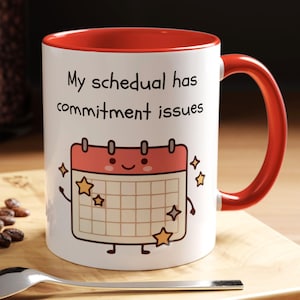 May include: White ceramic mug with a red interior and handle. The mug features the text "My schedual has commitment issues" and a cartoon calendar with stars. A spoon and a pastry are in the foreground.