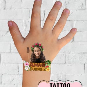 May include: A temporary tattoo featuring a cartoon image of a girl wearing a flower crown and holding a spear. The tattoo also includes a wooden sign with the text "Ana Turns 2" and tropical flowers and leaves. The tattoo is on a child's hand.