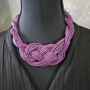 May include: A close-up of a purple beaded necklace. The necklace is made of many strands of small, round beads woven together in a braided design. The necklace is displayed on a black mannequin.