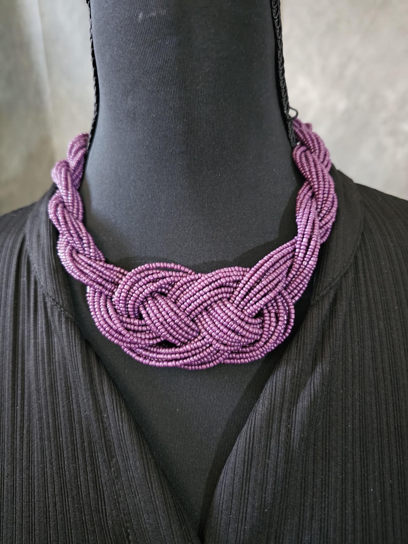 May include: A close-up of a purple beaded necklace. The necklace is made of many strands of small, round beads woven together in a braided design. The necklace is displayed on a black mannequin.