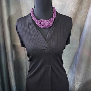 May include: A black dress with a V-neck is displayed on a mannequin. A vibrant purple beaded necklace is draped around the neck. The necklace is a chunky, braided design. The background is a muted gray and green.