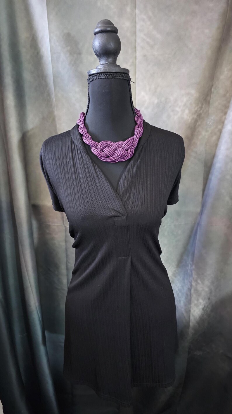 May include: A black dress with a V-neck is displayed on a mannequin. A vibrant purple beaded necklace is draped around the neck. The necklace is a chunky, braided design. The background is a muted gray and green.