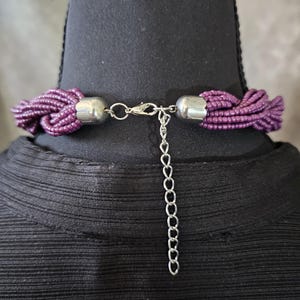 May include: A close-up of a purple beaded necklace with a silver clasp and extender chain. The necklace is displayed on a black mannequin. The beads are a deep purple color.