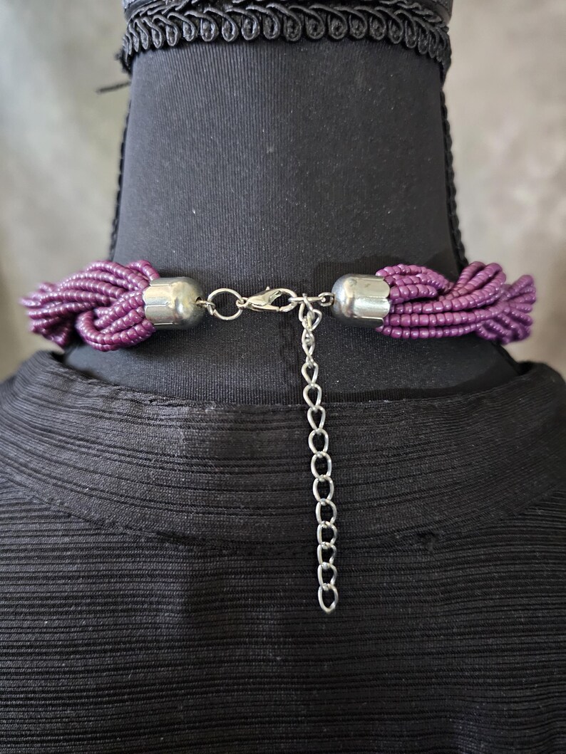 May include: A close-up of a purple beaded necklace with a silver clasp and extender chain. The necklace is displayed on a black mannequin. The beads are a deep purple color.