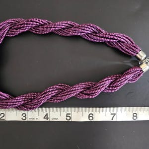 May include: A braided purple necklace with a silver-tone chain clasp. The necklace is approximately 10 inches long, with a knotted design at the center. The beads are a deep purple color.