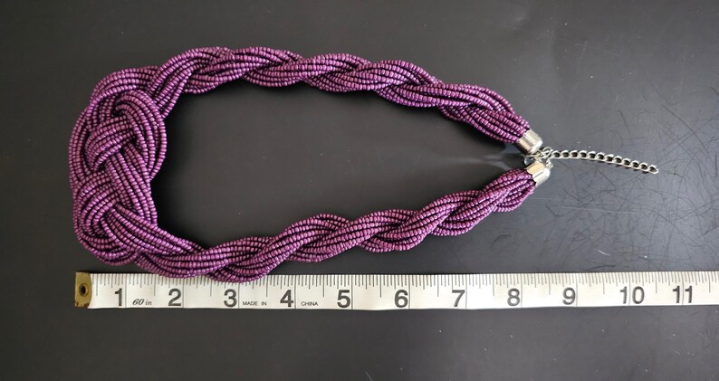 May include: A braided purple necklace with a silver-tone chain clasp. The necklace is approximately 10 inches long, with a knotted design at the center. The beads are a deep purple color.