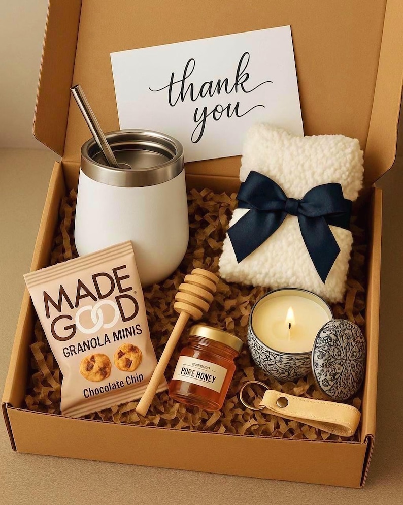 May include: A gift box filled with a variety of items, including a white thermal mug with a stainless steel lid and a straw, a small jar of honey, a candle in a white ceramic jar with a blue floral design, a packet of chocolate chip granola minis, a white fluffy pillow with a blue bow, and a small wooden honey dipper. The box is lined with brown paper shreds and includes a handwritten card that says "thank you".