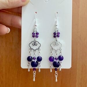 Puede incluir: Un par de pendientes colgantes con ganchos plateados. Cada pendiente presenta pequeñas cuentas moradas, un dije en forma de nube y cuentas colgantes en tonos morados y azul oscuro. Los pendientes se exhiben en una tarjeta blanca.