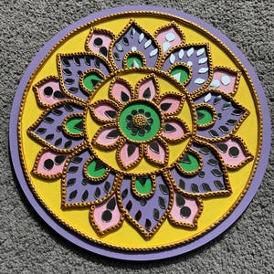 May include: A round, decorative mandala in shades of yellow, purple, pink, green, and gold. The design features a central flower shape surrounded by mirrored petal-like elements. The mandala is painted on a circular wooden base.