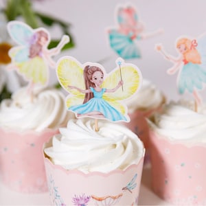May include: Cupcakes with white frosting and fairy-themed toppers. The fairy toppers feature watercolor illustrations of fairies with butterfly wings, in shades of blue, yellow, and pink. The cupcake liners are pink with floral and dragonfly designs.