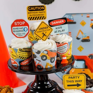May include: A black cake stand holds cupcakes with white frosting and construction-themed wrappers. The wrappers feature images of trucks and construction signs. Decorative signs read "CAUTION," "STOP," "DANGER," and "PARTY THIS WAY."