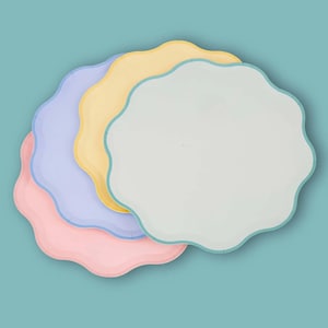 May include: Four pastel-coloured paper plates with a scalloped edge. The plates are in shades of pink, lavender, yellow, and light green, with a matching border. The plates are arranged on a teal background.