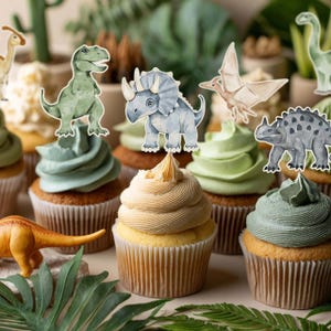 May include: A collection of cupcakes with various frosting colors, topped with dinosaur-themed decorations. The cupcakes have light brown wrappers and are arranged on a surface with green foliage. The dinosaur toppers include a T-Rex, Triceratops, and other prehistoric creatures.
