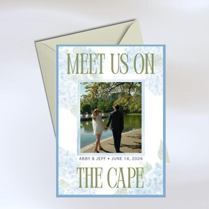 May include: A rectangular wedding invitation card with a light blue border and a floral pattern. The text reads MEET US ON THE CAPE and ABBY & JEFF JUNE 14, 2026. A central photograph shows two people walking along a path by a lake. A light envelope is behind.