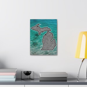 Op de afbeelding: Een canvas print met de omtrek van de staat Michigan, gevuld met een gedetailleerd zwart-wit patroon. De achtergrond toont een levendige turquoise en blauwe water scène, wat een opvallend contrast creëert. Het kunstwerk is op een wit oppervlak gemonteerd.