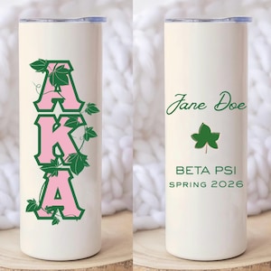 May include: A white cylindrical tumbler featuring the pink and green letters AKA intertwined with green ivy vines on one side. The other side displays the name Jane Doe in green script, a green ivy leaf, and the text BETA PSI SPRING 2026.