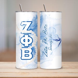 Può includere: Un bicchiere in acciaio inossidabile con coperchio e cannuccia. Il bicchiere presenta le lettere greche ZΦB in blu, con un design a collana di perle. L'altro lato ha il testo "Zeta Phi Beta Sorority, Inc" in blu, con un design astratto blu.