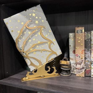 以下が含まれることがあります： グレーの表紙と金の星模様の書籍を支える、金色の金属製ブックスタンド。スタンドは装飾的な翼のデザインです。棚には他の本も見えます。